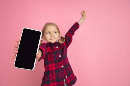 Showing blank screen. Caucasian little girls portrait on pink studio background. Beautiful female model with blonde hair. Concept of human emotions, facial expression, sales, ad, youth, childhood.の写真素材