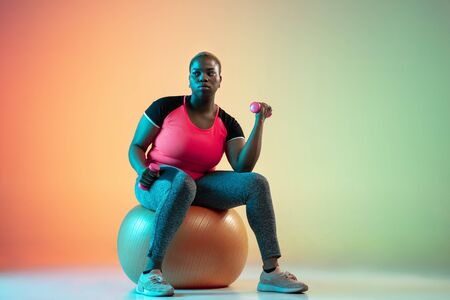 Young african-american plus size female models training on gradient background in neon light. Doing...