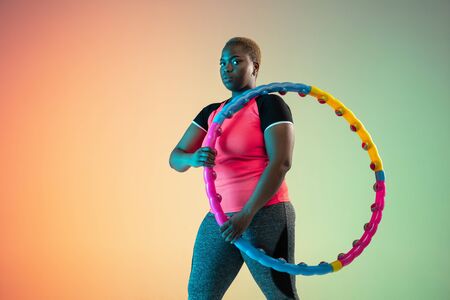 Young african-american plus size female models training on gradient background in neon light. Doing workout exercises with hoop. Concept of sport, healthy lifestyle, body positive, equality.の写真素材