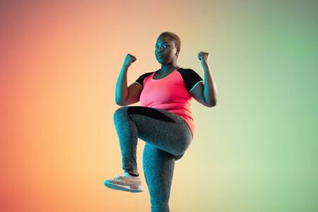Young african-american plus size female models training on gradient background in neon light. Doing...