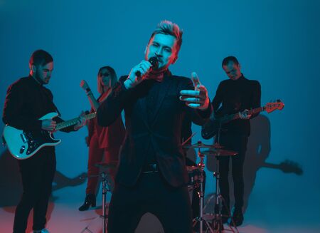 Young caucasian musicians, band performing on blue studio background in neon light. Concept of music, hobby, festival. Colorful portrait of modern artist. Attented and inspired. Art, cover band.の写真素材