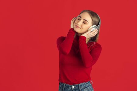 Listen to music. Monochrome portrait of young caucasian blonde woman isolated on red studio background. Beautiful model in shirt. Human emotions, facial expression, sales, ad concept. Youth culture.の写真素材