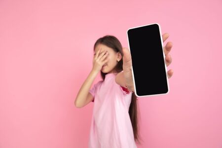 Shocked shows gadgets screen. Caucasian little girl portrait on pink studio background. Cute brunette model in shirt. Concept of human emotions, facial expression, sales, ad, childhood. Copyspace.の写真素材