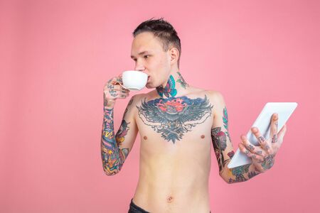 Drinks coffee, using tablet. Portrait of young man with freaky appearance on pink background. Unusual look with huge tattooes. Human emotions, facial expression, sales, ad concept. Youth culture.の写真素材