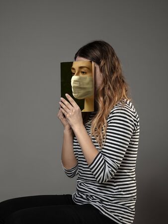 Happy world book day 2020, be safe and read to become someone else - woman covering face with book in mask while reading on grey studio background. Celebrating, education, art, protection concept.の写真素材