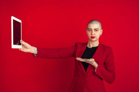 Taking selfie or vlog. Portrait of young caucasian bald woman isolated on red studio background. Beautiful female model in jacket. Human emotions, facial expression, sales, ad concept. Freaky culture.の写真素材
