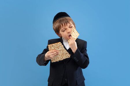 Joyfull eating matzah. Portrait of a young orthodox jewish boy isolated on blue studio background. Purim, business, festival, holiday, childhood, celebration Pesach or Passover, judaism, religion concept.の写真素材