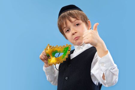 Trying carnival mask on. Portrait of a young orthodox jewish boy isolated on blue studio background. Purim, business, festival, childhood, celebration Pesach or Passover, judaism, religion concept.の写真素材