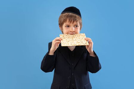 Joyfull eating matzah. Portrait of a young orthodox jewish boy isolated on blue studio background. Purim, business, festival, holiday, childhood, celebration Pesach or Passover, judaism, religion concept.の写真素材
