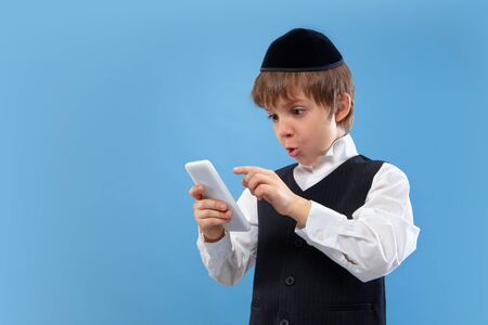 Shocked using phone. Portrait of a young orthodox jewish boy isolated on blue studio background....