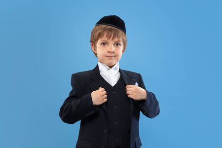 Posing confident, cute. Portrait of a young orthodox jewish boy isolated on blue studio background....