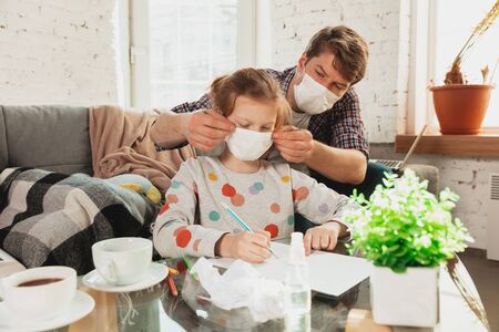 Father and daughter in face masks and gloves isolated at home with coronavirus respiratory symptoms such as fever, headache, cough in mild condition. Healthcare, medicine, quarantine, treatment concept.の写真素材