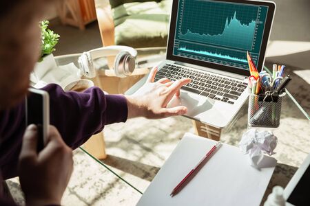 Checking graphs. Man working from home during coronavirus or COVID-19 quarantine, remote office concept. Young businessman, manager doing tasks with smartphone, laptop, tablet has online conference.の写真素材