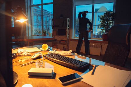 Need a rest. Man working in office alone during coronavirus or COVID-19 quarantine, staying to late night. Young businessman, manager doing tasks with smartphone, laptop, tablet in empty workspace.の写真素材