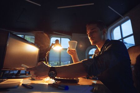 Attented. Man working in office alone during coronavirus or COVID-19 quarantine, staying to late night. Young businessman, manager doing tasks with smartphone, laptop, tablet in empty workspace.の写真素材