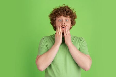 Shocked, astonished, wondered. Caucasian young mans monochrome portrait on green studio background. male curly model in casual style. Concept of human emotions, facial expression, sales, ad.の写真素材