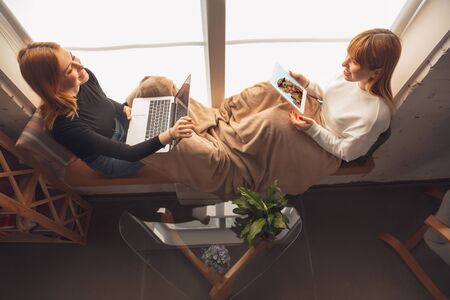 Best memories. Young friends, women using gadgets to watch cinema, photos, online courses, taking selfie or vlog. Two caucasian female models at home near window using laptop, tablet, smartphone.の写真素材