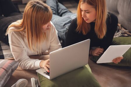 Education time. Young friends, women using gadgets to watch cinema, photos, online courses, taking selfie or vlog, shopping. Two caucasian girls at home using laptop, tablet, smartphone, headphones.の写真素材