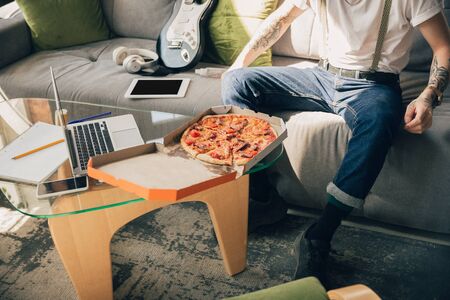 Eating pizza. Man studying at home during online courses, smart school. Getting classes or profession while isolated, quarantine against coronavirus spreading. Using laptop, smartphone, headphones.の写真素材