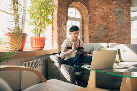 Man studying at home during online courses or free information by hisself. Becomes musician, guitarist while isolated, quarantine against coronavirus spreading. Using laptop, smartphone, headphones.の写真素材
