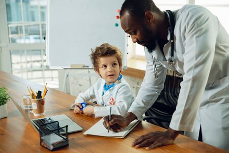 Caucasian teenboy as a doctor consulting for patient, giving recommendation, treating. Little doctor during working with older colleague. Concept of childhood, human emotions, health, medicine.の写真素材