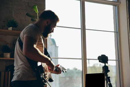 Caucasian musician playing guitar during online concert at home isolated and quarantined. Using camera, laptop, streaming, recording courses. Concept of art, support, music, hobby, education.の写真素材