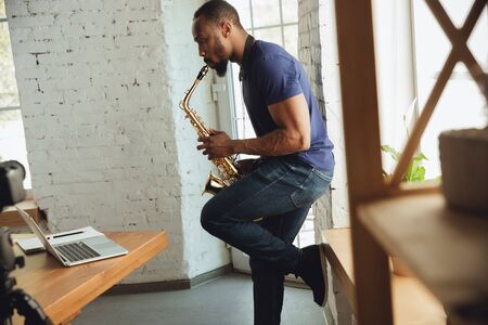 African-american musician playing saxophone during online concert at home isolated and quarantined. Using camera, laptop, streaming, recording courses. Concept of art, support, music, hobby, education.の写真素材