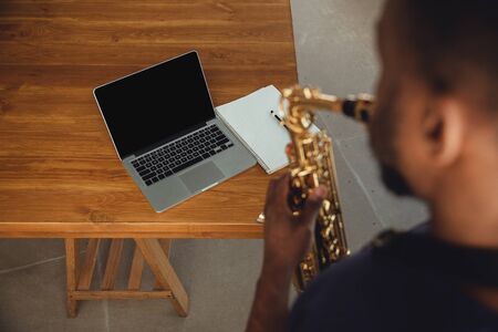 African-american musician playing saxophone during online concert at home isolated and quarantined. Using camera, laptop, streaming, recording courses. Concept of art, support, music, hobby, education.の写真素材