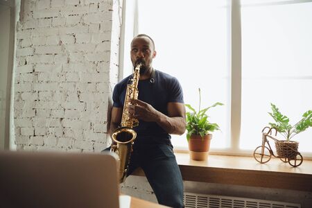 African-american musician playing saxophone during online concert at home isolated and quarantined. Using camera, laptop, streaming, recording courses. Concept of art, support, music, hobby, education.の写真素材
