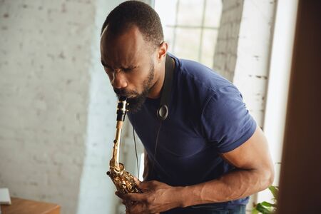 African-american musician playing saxophone during online concert at home isolated and quarantined. Using camera, laptop, streaming, recording courses. Concept of art, support, music, hobby, education.の写真素材