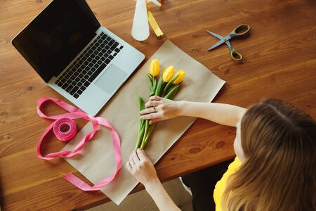 Florist at work: woman shows how to make bouquet with tulips. Young caucasian woman gives online workshop of doing gift, present for celebration. Working at home while isolated, quarantined concept.の写真素材