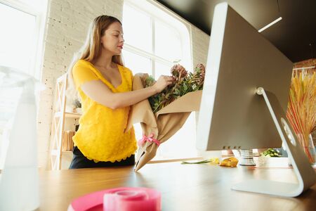Florist at work: woman shows how to make bouquet. Young caucasian woman gives online workshop of doing gift, present for celebration, with computer. Working at home while isolated, quarantined concept.の写真素材