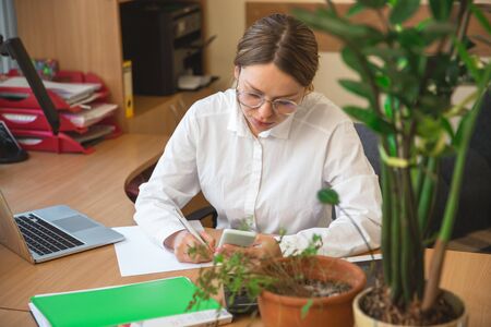 Using gadgets. Caucasian entrepreneur, businesswoman, manager working concentrated in office. Looks serios and busy, wearing classic attire. Concept of work, finance, business, success, leadership.の写真素材