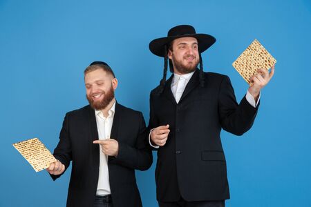 Eating matzah, inviting. Portrait of a young orthodox jewish men isolated on blue studio background....