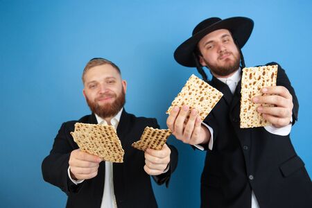 Eating matzah, inviting. Portrait of a young orthodox jewish men isolated on blue studio background. Purim, business, festival, holiday, celebration Pesach or Passover, judaism, religion concept.の写真素材
