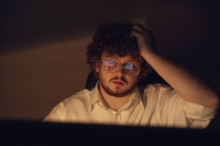 Focused and attented. Man working in office alone during COVID-19 quarantine, staying to late night. Young businessman, manager doing tasks with smartphone, laptop, tablet in empty workspace.の写真素材