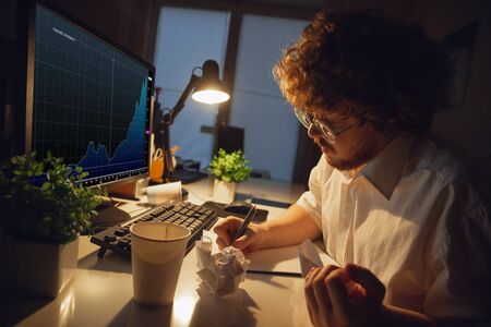 Making notes, paperwork. Man working in office alone during COVID-19 quarantine, staying to late night. Young businessman, manager doing tasks with smartphone, laptop, tablet in empty workspace.の写真素材