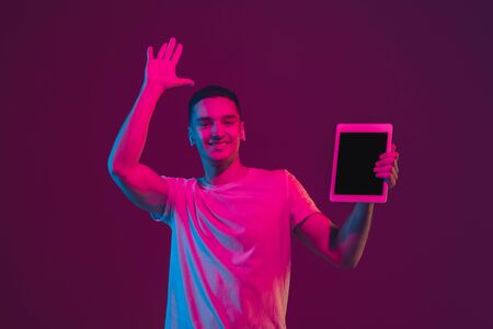 Showing blank screen. Caucasian mans portrait isolated on pink-purple studio background in neon light. Male model with devices. Concept of human emotions, facial expression, sales, ad. Copyspace.の写真素材