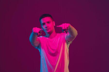 Pointing, choosing. Caucasian mans portrait isolated on pink-purple studio background in neon light. Beautiful male model with earphones. Concept of human emotions, facial expression, sales, ad.の写真素材