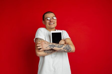 Blank tablet screen. Caucasian young mans portrait isolated on red studio background. Beautiful male model with devices, gadgets. Concept of human emotions, facial expression, sales, ad, technologies.の写真素材
