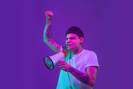 Shouting with speaker. Caucasian mans portrait isolated on purple studio background in pink neon light. Beautiful male model in casual. Concept of human emotions, facial expression, sales, ad.の写真素材
