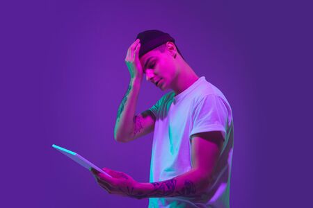Upset using tablet. Caucasian mans portrait isolated on purple studio background in pink neon light. Beautiful male model in casual. Concept of human emotions, facial expression, sales, ad.の写真素材