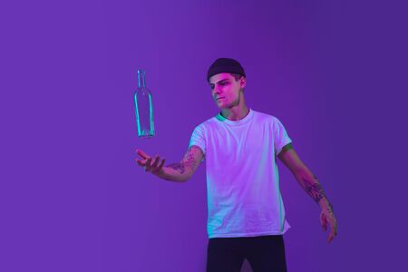 Throws up a bottle of water. Caucasian mans portrait isolated on purple studio background in pink neon light. Beautiful male model in casual. Concept of human emotions, facial expression, sales, ad.の写真素材