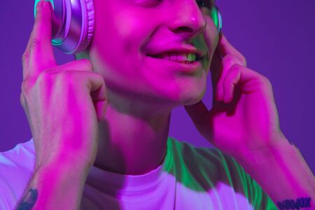 Listen to music, close up. Caucasian mans portrait isolated on purple studio background in pink neon light. Beautiful male model in casual. Concept of human emotions, facial expression, sales, ad.の写真素材