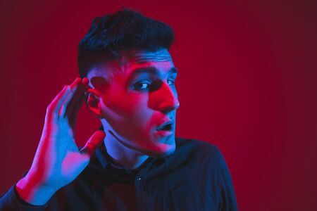 Listening to secrets with hand on ear. Caucasian young mans portrait isolated on red studio...