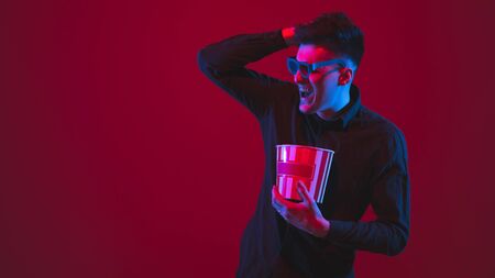 Looking throught 3D-eyewear. Caucasian young mans portrait isolated on red studio background in neon light. Beautiful model. Concept of human emotions, facial expression, sales, ad, youth, devices.の写真素材