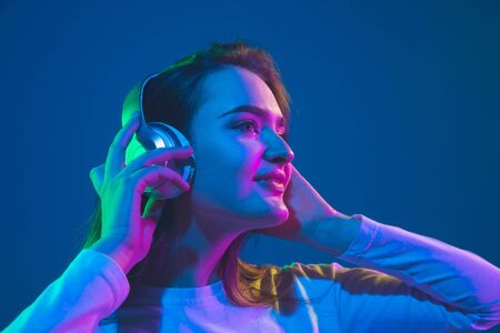 Listening to music. Caucasian young womans portrait isolated on blue studio background in neon light. Beautiful female model. Concept of human emotions, facial expression, sales, ad, youth culture.の写真素材