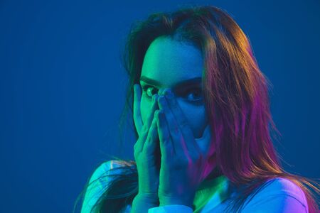 Hiding face with hands. Caucasian young womans portrait isolated on blue studio background in neon light. Beautiful female model. Concept of human emotions, facial expression, sales, ad, youth culture.の写真素材