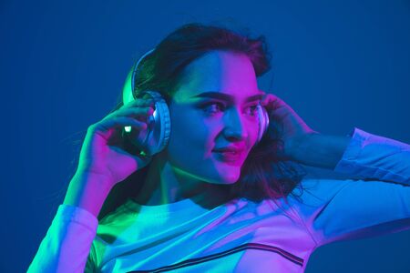 Listening to music. Caucasian young womans portrait isolated on blue studio background in neon light. Beautiful female model. Concept of human emotions, facial expression, sales, ad, youth culture.の写真素材