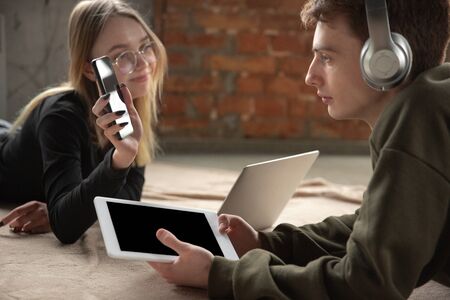 Attractive young couple using devices together, tablet, laptop, smartphone, headphones wireless. Communication, gadgets concept. Technologies connecting people in self-insulation. Lifestyle at home.の写真素材